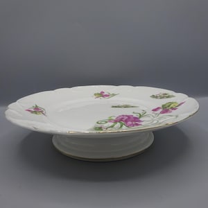 May include: White porcelain cake stand with a scalloped edge and floral design. The flowers are pink and green. The stand has a gold rim and a pedestal base.