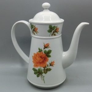 Könnte beinhalten: Weiße Kaffeekanne aus Keramik mit Deckel und Henkel. Die Kanne hat ein Blumenmuster mit orangen Rosen und grünen Blättern.