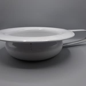 Puede incluir: Orinal de cerámica blanca, con un borde ancho y plano y un asa curvada. El orinal tiene una base redondeada y un acabado liso y brillante. El asa está unida al lateral y se curva hacia arriba.