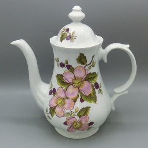 Könnte beinhalten: Weiße Porzellan-Kaffeekanne mit floralem Design mit rosa und grünen Blumen. Der Deckel hat ein florales Design mit violetten Beeren.
