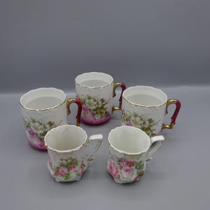 Può includere: Cinque tazze e tazze antiche in porcellana. Le tazze presentano disegni floreali in rosa e bianco, con finiture dorate e manici rossi. Le tazze hanno un effetto sfumato rosa alla base. Un set classico per tè o caffè.
