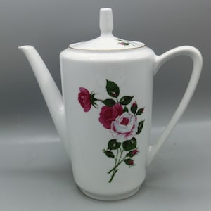 Op de afbeelding: Witte keramische koffiepot met een lange tuit, handvat en deksel. De pot is versierd met een bloemenontwerp met roze en rode rozen met groene bladeren. De pot staat op een lichtgrijze achtergrond.