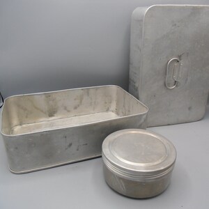 May include: A set of three vintage aluminum containers. Includes a rectangular box, a tall rectangular box with a handle, and a small round container with a lid. The containers are all a silver color.