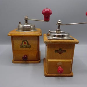 Peut inclure: Deux moulins à café en bois vintage avec des poignées et des boutons rouges. Les moulins ont un corps en bois brun clair, un dessus en métal argenté et un petit tiroir. Chaque moulin a un logo à l'avant.