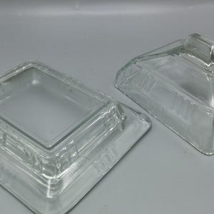 Puede incluir: Un mantequillero de vidrio transparente con tapa. El mantequillero tiene una base cuadrada y una parte superior rectangular. La tapa tiene una pequeña manija en la parte superior.