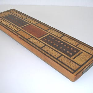May include: Wooden cribbage board with a rectangular shape. The board features a light brown base with dark brown and reddish-brown inlaid sections. It has multiple rows of holes for pegs and a metal clasp.