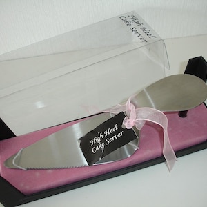 May include: A stainless steel cake server in the shape of a high heel shoe, presented in a black box with pink tissue paper. The server has a serrated edge and is tied with a pink ribbon. The text "High Heel Cake Server" is on a black tag.