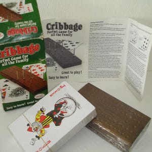 May include: A cribbage set featuring a wooden board, pegs, playing cards, and instructions. The box is labelled "Cribbage, Perfect Game for all the Family." A deck of cards with a joker is visible. A classic family board game.