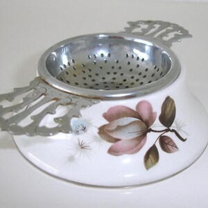 May include: A white ceramic tea strainer stand featuring a painted pink flower with green leaves and brown stems. The stand has a circular opening designed to hold a silver-toned metal tea strainer with decorative scalloped handles on each side.