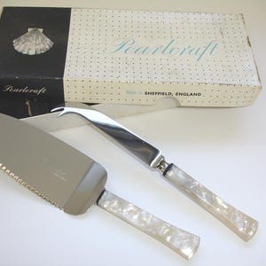 May include: A silver-toned cake server and a cheese knife with white pearlescent handles. The items are presented with a rectangular box labeled Pearlcraft, Made in SHEFFIELD, ENGLAND, featuring a black and white dotted pattern and a shell graphic.