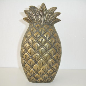 May include: A metallic gold-toned pineapple sculpture featuring a textured, repeating diamond-shaped pattern across the body and a detailed, pointed leaf crown at the top. The object has a weathered, antique finish with dark recessed areas.