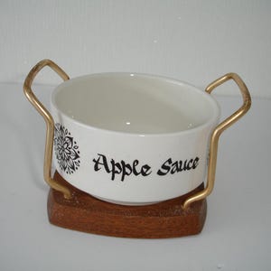 May include: White ceramic bowl with a black and white floral design and the words "Apple Sauce" on the side. The bowl is on a gold metal stand with a wooden base.