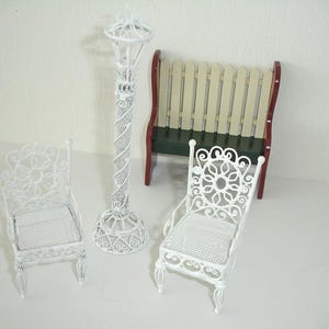 May include: A set of miniature white metal garden furniture, comprising two chairs, a lamppost, and a bench. The chairs have floral designs, and the lamppost has a spiral pattern. The bench has a red and green base with a white picket fence back.