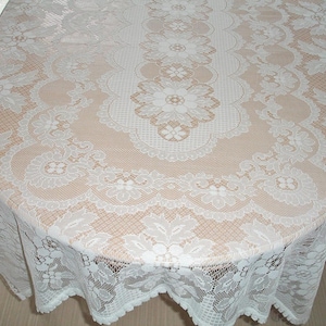 May include: A white lace tablecloth with a floral design covers a round table. The tablecloth has a scalloped edge and intricate patterns. The table is set on a light-colored wooden floor.