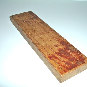 May include: A wooden cribbage board with a worn, reddish-brown finish. The board has numerous holes for pegs, and the edges are slightly rounded.