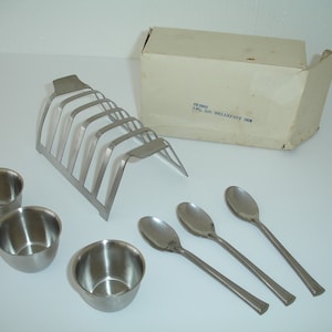 May include: A stainless steel breakfast set with a toast rack, three small bowls, and four spoons. The set comes in a white cardboard box with the text "75-2003 4PC. 50% BREAKFAST SET" printed on it.