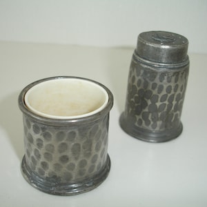 May include: Two antique metal containers with a hammered texture. One is cylindrical with a white interior, and the other is a taller, tapered shape with a lid. The containers are likely for holding small items or serving as decorative pieces.
