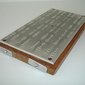 May include: A wooden cribbage board with a metal playing surface. The board has a brown wood frame and a silver metal playing surface with holes for pegs. The board has two metal corner pieces.