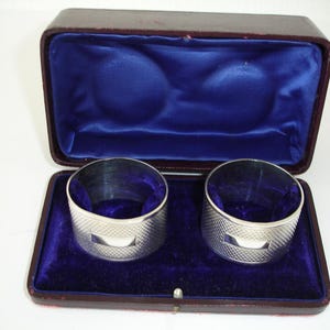 May include: Two silver napkin rings in a brown velvet lined box. The rings have a textured design and a small rectangular opening.