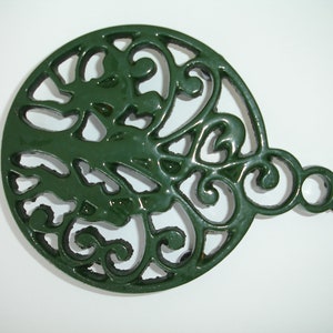 May include: Green cast iron trivet with a decorative floral design. The trivet has a handle on the side.