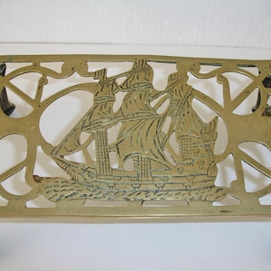 May include: A rectangular brass trivet featuring an openwork design of a three-masted sailing ship on the water. The metal stand has four legs with claw-style feet and a polished gold-toned finish with intricate cut-out patterns throughout.