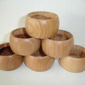 May include: A stack of six wooden napkin rings. Each ring is a light brown color and has a smooth, rounded shape. The rings are stacked in a slightly haphazard arrangement, with some rings partially obscuring others. The wood grain is visible.