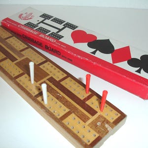 May include: A wooden cribbage board with pegs, alongside its red and white box. The board is made of light and dark wood, with white and red pegs inserted. The box features card suit symbols.