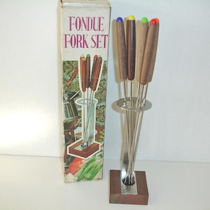 Peut inclure: Un ensemble de fourchettes à fondue vintage avec une boîte indiquant "FONDUE FORK SET". L'ensemble comprend six fourchettes avec des manches en bois et des embouts colorés, présentées dans un support en métal et en bois. La boîte a une illustration colorée.