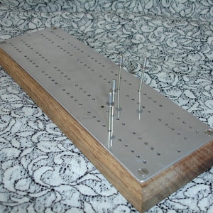 May include: A cribbage board made of wood and metal. The playing surface is metal, with many small holes for pegs. Several metal pegs are inserted. The wooden base has a natural finish. The board is rectangular.