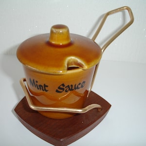 May include: A brown ceramic jar with a lid and a metal handle, labelled "Mint Sauce". The jar sits on a wooden base.