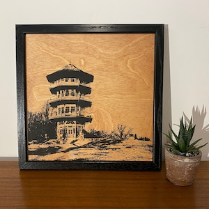 May include: A black and white silhouette of a pagoda-style building on a wood background. The building has multiple tiers and a pointed roof. The image is framed in a black frame.
