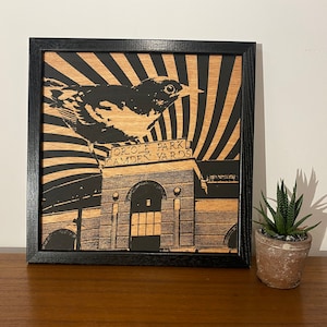 May include: A black framed wood print of a black and white illustration of a bird perched on a sign that reads "Oriole Park Camden Yards". The background is a black and white striped pattern.