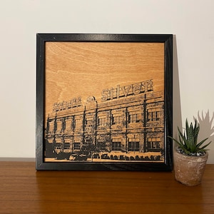 May include: A black framed wood sign with a laser-engraved image of a building with the text "Schlitz Silver" on the top.