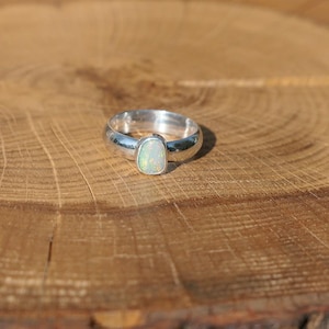 May include: A silver ring featuring a rectangular opal gemstone. The ring's band is thick and polished, and the opal displays a subtle iridescence. The ring is set on a wooden surface.