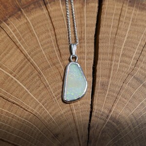 May include: A silver chain necklace with a white opal pendant. The opal is set in a silver bezel and has a teardrop shape. The necklace is photographed on a wooden surface.