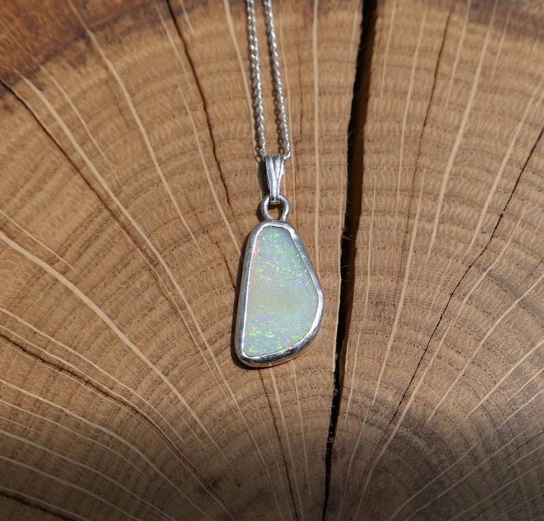 May include: A silver chain necklace with a white opal pendant. The opal is set in a silver bezel and has a teardrop shape. The necklace is photographed on a wooden surface.
