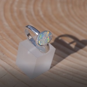 May include: A silver ring featuring an oval opal gemstone. The opal has a white base with flashes of green, blue, and orange.