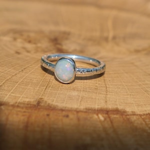 May include: A silver ring featuring a textured band and an oval opal gemstone. The opal exhibits a play of colour. The ring is displayed on a wooden surface, emphasizing the jewellery's design and craftsmanship.