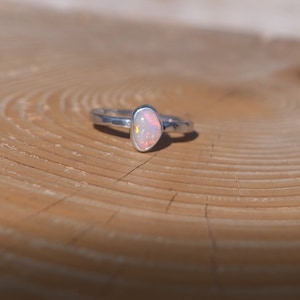 May include: A delicate silver ring featuring a small, pink opal gemstone. The ring is displayed on a wooden surface.