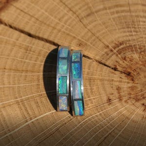 May include: Two silver rings with a row of iridescent opal stones set in a silver band. The rings are on a wooden surface.