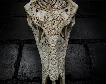 May include: A white deer skull with intricate carvings of flowers and a tree of life design. The skull is mounted on a black brick wall.