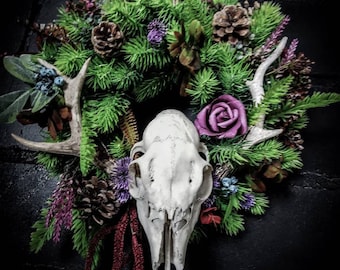 May include: A white deer skull with antlers is surrounded by a wreath of green pine branches, purple flowers, and pine cones.