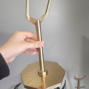 May include: A gold-coloured, geometric display stand with a hexagonal base and a vertical support that splits into two prongs at the top. The stand is held by a person's hand. Other decorative items are in the background.