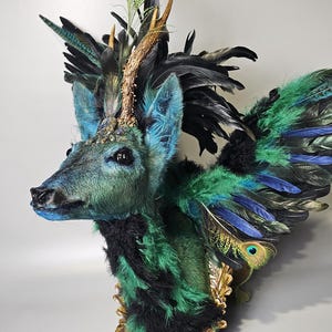 Puede incluir: Una cabeza de ciervo disecada con cuernos, adornada con plumas verdes, azules y negras. La piel del ciervo está pintada de un verde y azul vibrantes, y sus ojos son negros. La cabeza de ciervo está montada sobre una base dorada.