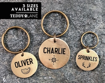 May include: Three gold-colored metal dog tags with key rings. The tags are engraved with the names "Oliver", "Charlie", and "Sprinkles". The Oliver tag has a watermelon slice design, the Charlie tag has a compass design, and the Sprinkles tag has a pair of antlers.