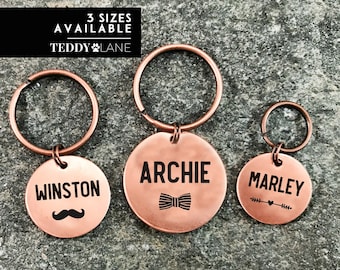 May include: Three copper dog tag keychains with different names and designs. The keychains are on a gray textured background. The text on the keychains reads "Winston", "Archie", and "Marley".