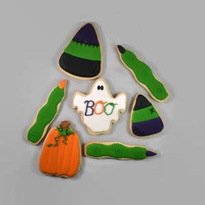 May include: Assortment of Halloween-themed sugar cookies. Includes a pumpkin, ghost with "BOO" text, candy corn, and green finger cookies. The cookies are decorated with orange, green, purple, black, and white icing.