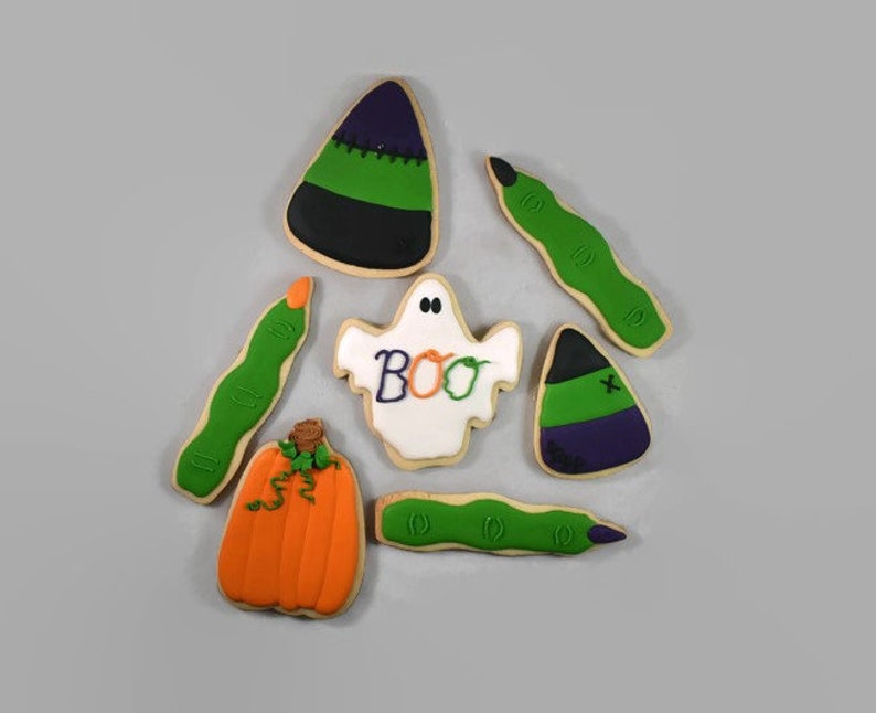 May include: Assortment of Halloween-themed sugar cookies. Includes a pumpkin, ghost with "BOO" text, candy corn, and green finger cookies. The cookies are decorated with orange, green, purple, black, and white icing.