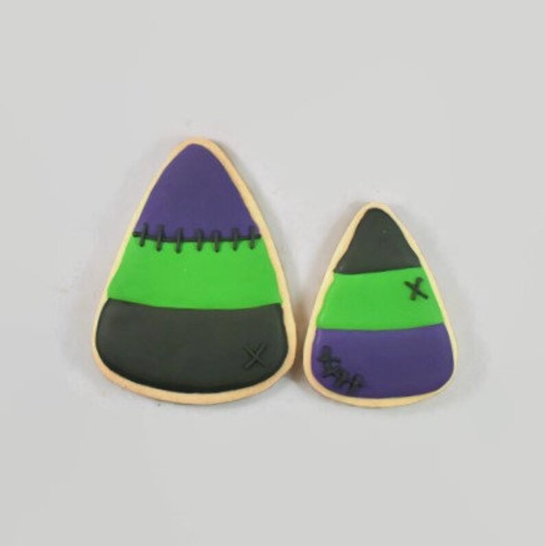 May include: Two Halloween-themed cookies shaped like candy corn. The larger cookie has a black base, a green middle, and a purple top with green stitching. The smaller cookie has a similar design.