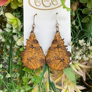Puede incluir: Pendientes de madera en forma de lágrima con diseño de libélulas y flores. Los pendientes son de color marrón claro con un patrón detallado cortado con láser. Los ganchos para las orejas son de color cobre. La tarjeta tiene el texto "Chi Chic Chi".